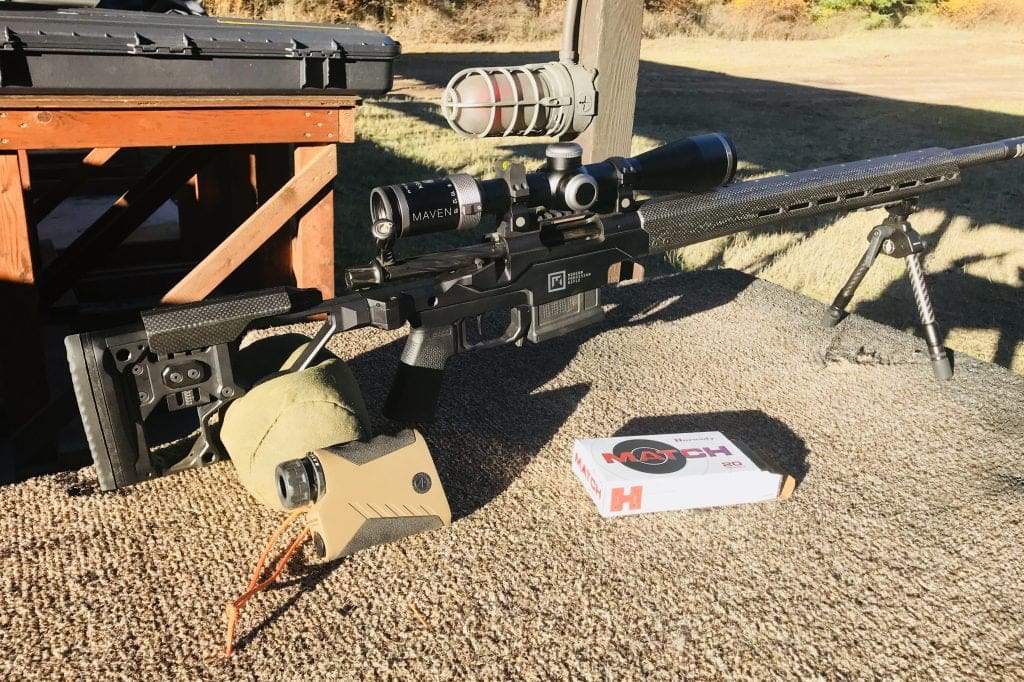 Picture of a scoped rifle sitting on a shooting bench with a rangefinder and box of bullets.